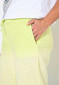 Lichtgele gaasbroek met een gestructureerde afwerking, een drawstring taille en zijzakken, afgebeeld terwijl het door een hand wordt vastgehouden.