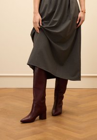 Burgundy leather knee-high boots with a pointed toe and block heel, paired with a flowing gray dress, set against a light wooden floor.