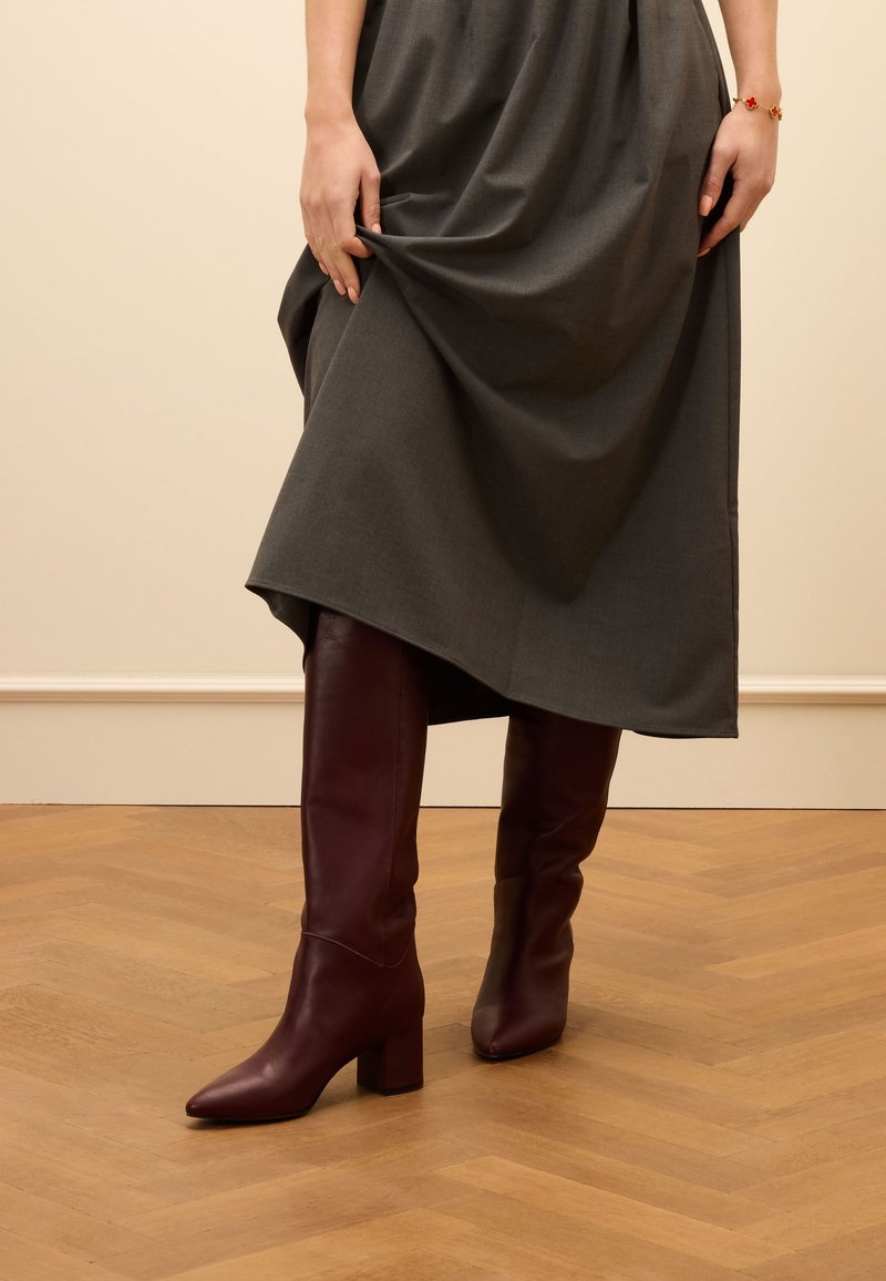 Burgundy leather knee-high boots with a pointed toe and block heel, paired with a flowing gray dress, set against a light wooden floor.