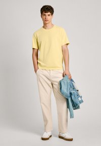Gul bomulds T-shirt, krämfarvede bukser, hvide sneakers med brune såler, holder en lys blå denimjakke, står foran en neutral baggrund.