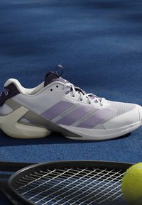Chaussure de sport blanche et violette à côté d'une raquette de tennis et d'une balle de tennis jaune sur une surface de court bleue.