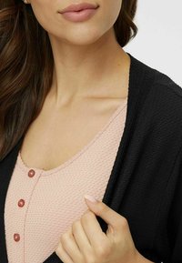 Svart cardigan över en ljusrosa texturerad topp med runda knappar. Tyget verkar mjukt, och designen är öppen fram, i avslappnad stil.