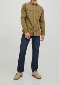 Khakifarbenes Button-up-Hemd mit zwei Brusttaschen, kombiniert mit dunkelblauen Jeans und beigen Schuhen. Das Hemd hat einen klassischen Kragen und eine taillierte Passform.