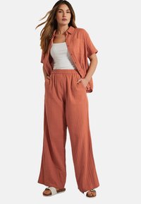Roodachtige, lichte, gewatteerde shirt en bijpassende wijde broek. Het heeft een knoopsluiting aan de voorkant en zijzakken. Gecombineerd met platte sandalen.