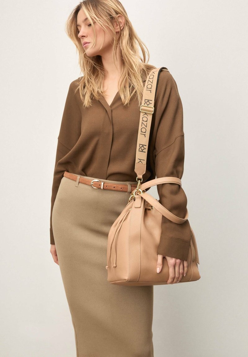 Naine pruunis pluusis ja beežis seelikus hoiab käes pruuni naha käekotti, millel on lai rihm ja "kazar" kaubamärgi logoga.