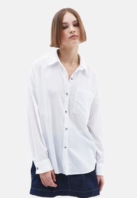 Witte lange mouwen button-up shirt met een kraag en een borstzak. Gladde textuur en zwarte knopen; gedragen met een donkere spijkerrok.