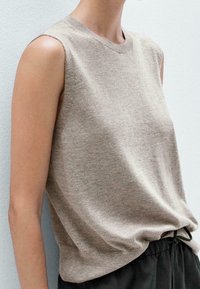 Beige ilmanhihaista neulepaitaa, jossa on pyöreä pääntie ja pehmeä rakenne, yhdistettynä tummiin kiristysnuorilla varustettuihin housuihin. Lähikuva rentosta istuvuudesta.