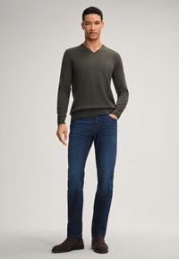 Pullover verde scuro a V abbinato a jeans blu. Il modello sta in piedi, con le mani in tasca, indossando stivali marroni. Sfondo minimalista.
