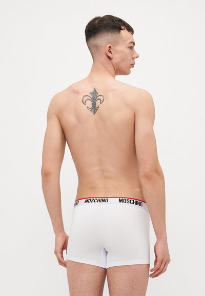White cotton boxer shorts with a black and red Moschino logo on the waistband; model has a tattoo on the back.