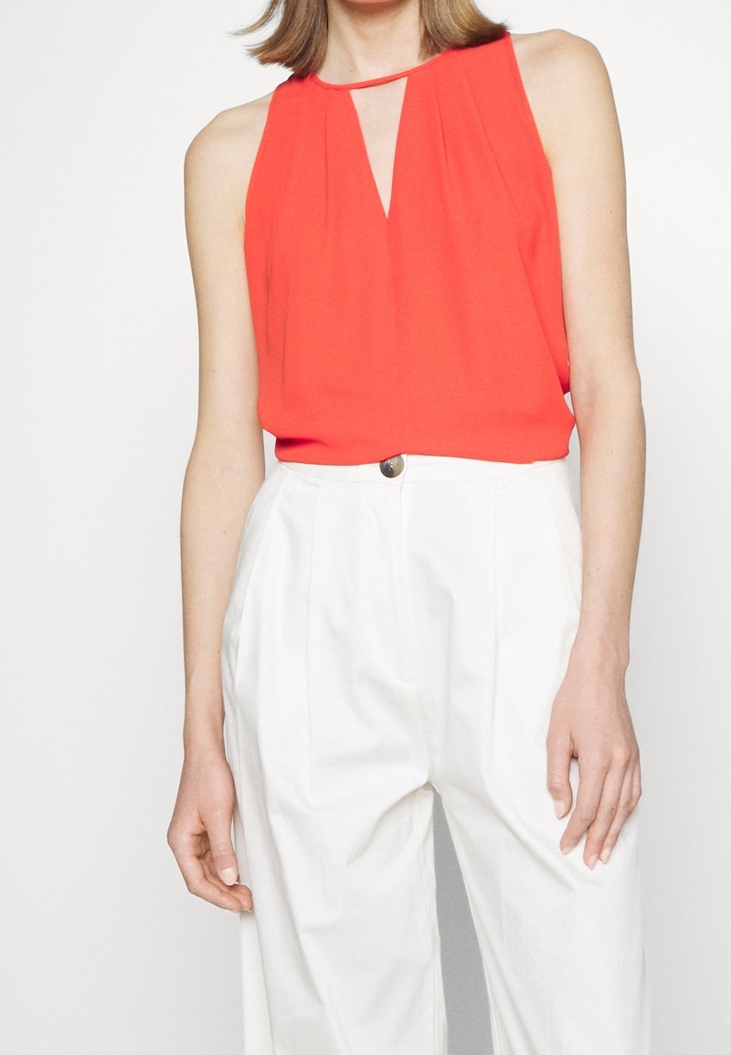 Femme portant un chemisier rouge sans manches avec une découpe à l'avant et un pantalon blanc taille haute avec fermeture à boutons, debout devant un fond uni.