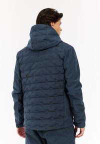 Man in een donkerblauwe gewatteerde jas met capuchon, gezien van achteren tegen een plain witte achtergrond.
