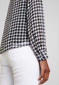 Blusa a motivi houndstooth bianco e nero con polsini a bottoni, realizzata in tessuto trasparente, abbinata a jeans di denim bianchi.