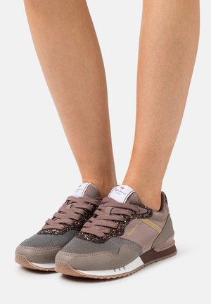 Jambes portant des baskets beiges et taupe avec des accents pailletés, en mesh, et des semelles en caoutchouc, debout sur une surface blanche.