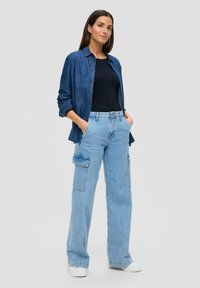 Lichtblauwe cargobroek met zijzakken, gecombineerd met een donkerblauw aansluitend topje en een donkerdenim knoopshirt. Model staat casual.
