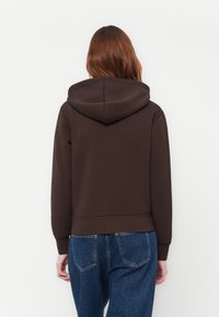 Persoon met lang bruin haar, gekleed in een donkerbruine hoodie en blauwe spijkerbroek, met de rug naar de kijker gekeerd tegen een effen lichte achtergrond.