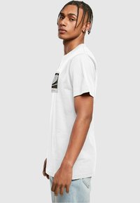 Hvid bomulds T-shirt med korte ærmer, med et sort og hvidt grafisk print på forsiden. Model iført lyseblå jeans.