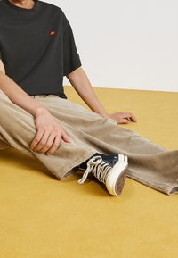 T-shirt noir à manches courtes avec un petit logo coloré, pantalon en velours côtelé beige, et baskets montantes bleu marine sur une surface jaune.