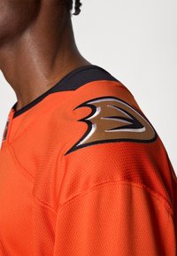 Oranž sportlik särg, millel on võrgusilma tekstuur, must kaelus ja seljal silmapaistev pruuni ja valge logoga emblem.