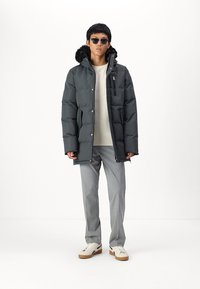 Doudoune noire avec capuche bordée de fourrure, poche zippée sur la poitrine, associée à un pantalon gris clair et des baskets blanches, accessoirisée de lunettes de soleil.