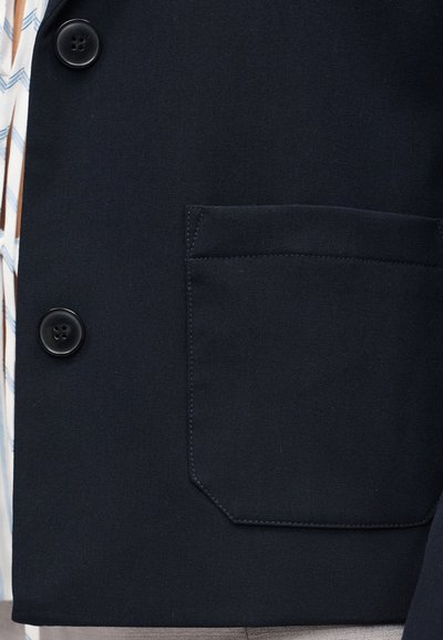 Σκούρο μπλε σακάκι με δύο μαύρα κουμπιά, μια μεγάλη τσέπη μπροστά, φορεμένο πάνω από λευκό πουκάμισο με μπλε ρίγες σε σχήμα βέλους.