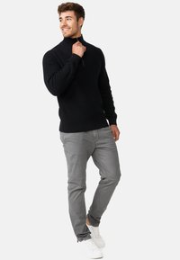 Pull en maille noir avec col haut et zip, associé à un pantalon gris et des baskets blanches. Le modèle pose avec une main sur le col.