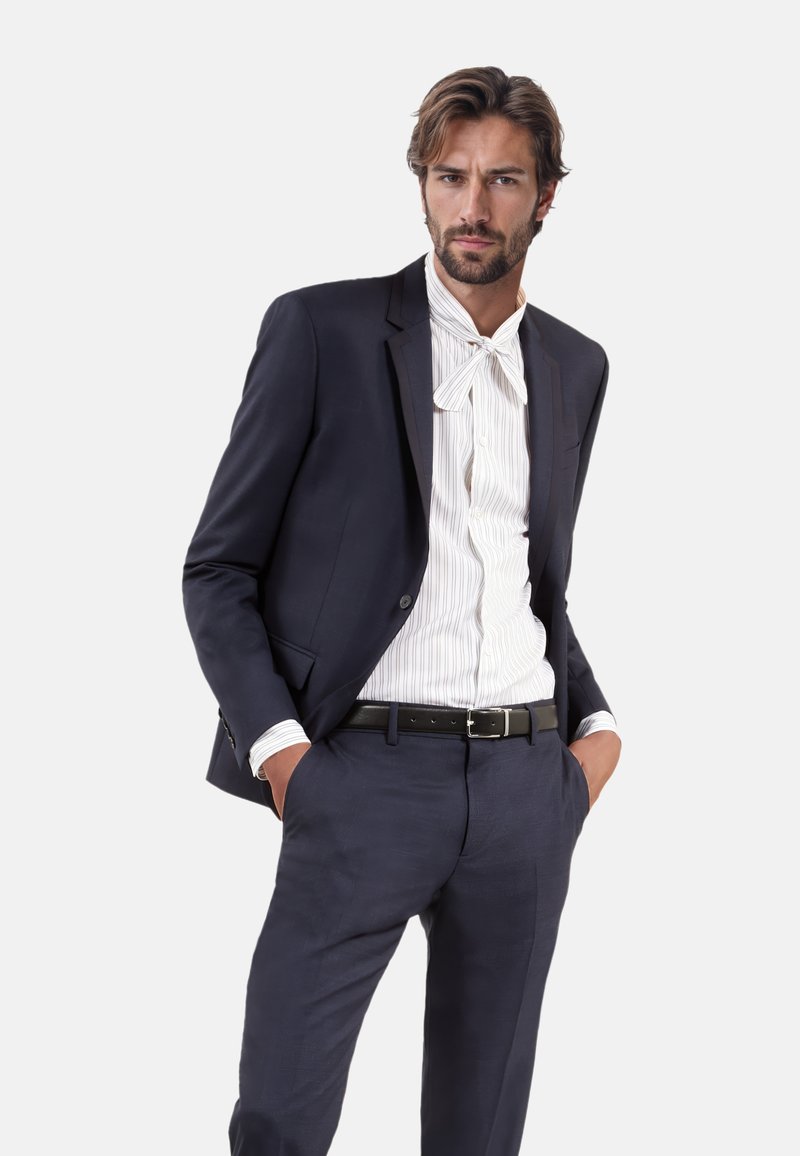 Blazer blu navy con pantaloni abbinati, abbinato a una camicia bianca a righe sottili. Presenta un papillon e una cintura nera con una fibbia argentata.