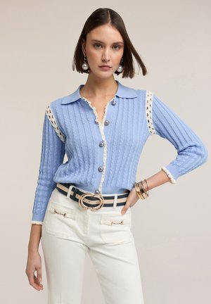 Kvinde iført en lyseblå strikket cardigan med hvide blonderdetaljer, hvide bukser, et sort og hvidt bælte og store perleøreringe, poserer med hånden på hoften.