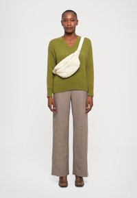 Olivgrüner Strickpullover mit V-Ausschnitt, beiger Kunstpelz-Umhängetasche, braune karierte weit geschnittene Hose und braune Stiefeletten.