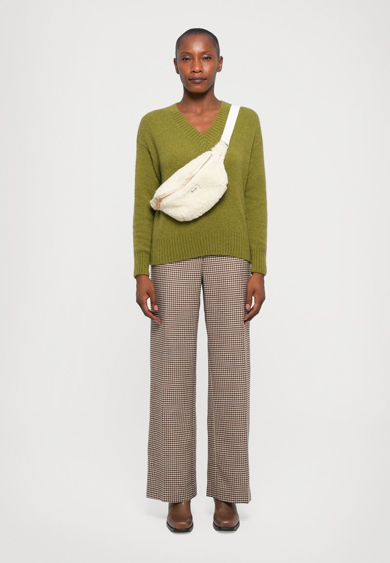Olivgrüner Strickpullover mit V-Ausschnitt, beiger Kunstpelz-Umhängetasche, braune karierte weit geschnittene Hose und braune Stiefeletten.