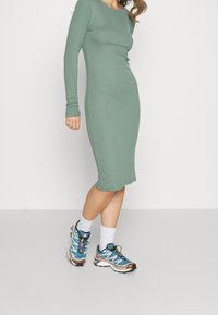 Abito midi verde a maniche lunghe con silhouette aderente. Abbinato a scarpe sportive blu con accenti bianchi e marroni e calzini bianchi.