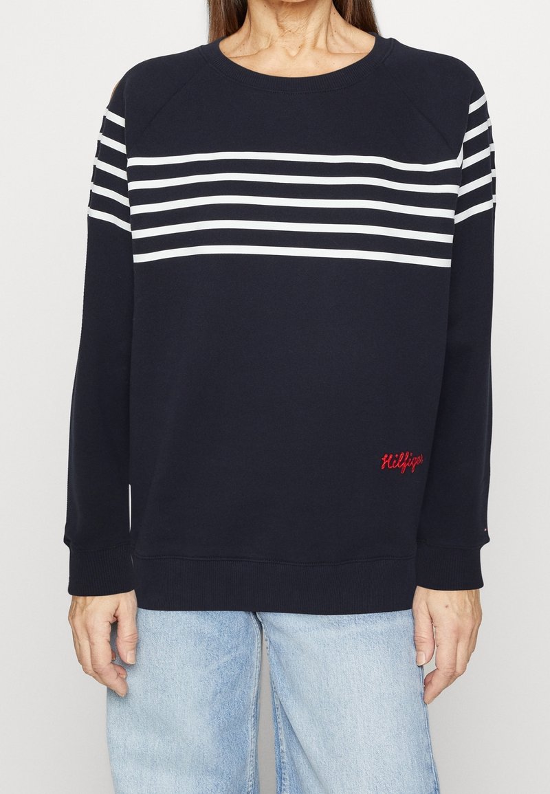 Femme portant un pull bleu marine avec des rayures blanches horizontales sur la poitrine et le haut des manches, associé à un jean bleu clair.