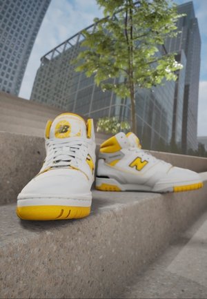 Hvide og gule New Balance-sneakers placeret på betontrapper med moderne glasbygninger og et træ i baggrunden.