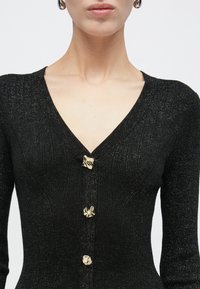 Μαύρο ριμπ ζακέτα με V-λαιμόκοψη, μακριά μανίκια και τρία ακανόνιστα χρυσά κουμπιά μπροστά, φορεμένη από άτομο.