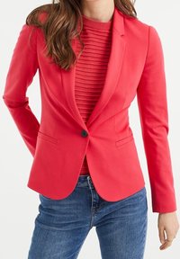 Korálový blazer s úzkým střihem, jedním knoflíkem na zapínání a slim fit, spárovaný s strukturovaným červeným topem a modrými džíny.