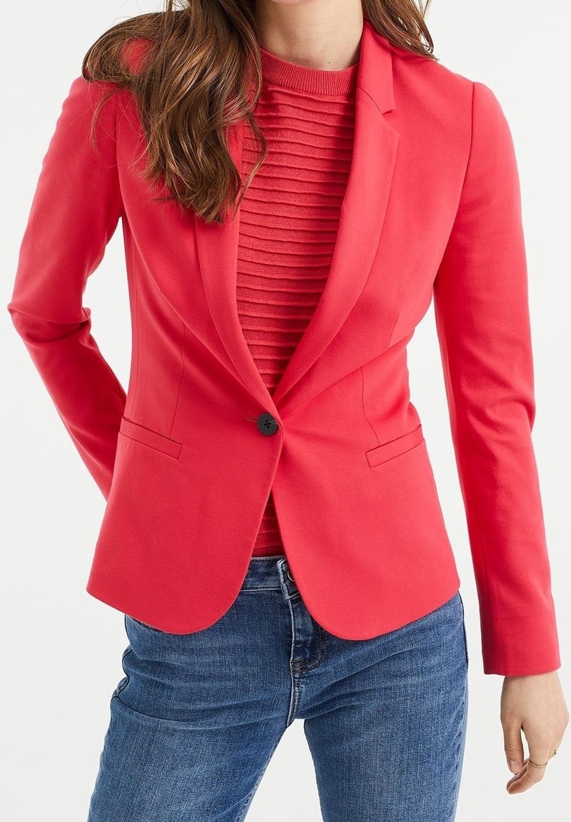 Korálový blazer s úzkým střihem, jedním knoflíkem na zapínání a slim fit, spárovaný s strukturovaným červeným topem a modrými džíny.