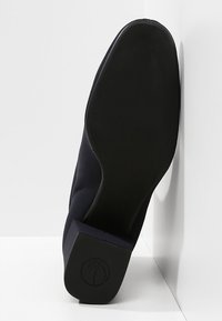 Semelle noire d'une pantoufle ou chaussure relevée contre un mur d'angle blanc, montrant un dessous lisse et un petit talon carré avec un logo en relief.