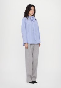 Camicia a righe blu e bianche con dettagli a colletto a forma di rosa, abbinata a pantaloni larghi grigi chiari e scarpe nere a punta.