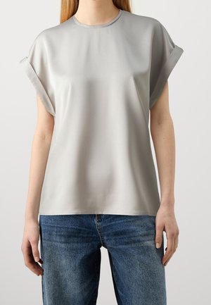 Femme portant un chemisier à manches courtes gris clair avec des revers retroussés, associé à un jean en denim bleu, debout devant un arrière-plan uni.