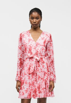 Mujer vistiendo un vestido de manga larga con estampado floral en tonos rosa y rojo, con botones en la parte delantera y un cinturón anudado en la cintura, posando frente a un fondo liso.