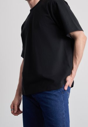 Schwarzes kurzärmliges T-Shirt mit lockerer Passform und rundem Halsausschnitt, aus glattem Stoff gefertigt. Besitzt ein kleines, dezentes Logo auf der Brust.