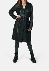 Abrigo trench de cuero negro con cuello de solapa, cintura ceñida con cinturón y mangas largas. Combinado con leggings grises acanalados y botines negros hasta el tobillo.