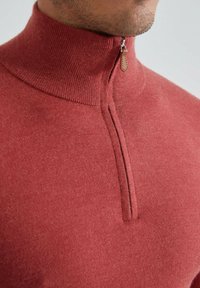 Pull rouge texturé avec un col haut et une fermeture zippée à mi-hauteur, fabriqué à partir d'un matériau doux avec une couture propre au niveau du col.