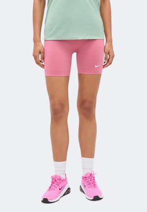 Άτομο που φοράει ροζ ποδηλατικά σορτς Nike, λευκές κάλτσες και ροζ αθλητικά παπούτσια Nike, στέκεται με τα χέρια χαλαρά στα πλάγια σε απλό φόντο.
