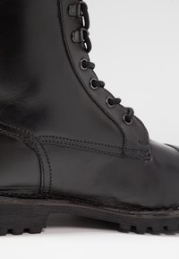 Botte en cuir noir à tige haute, avec une finition lisse, un laçage à œillets et une semelle en caoutchouc robuste avec motifs de crantage visibles.