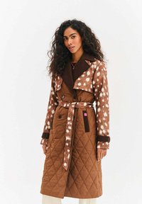 Brązowy pikowany płaszcz trencz z brązowo-kremowymi łatami z futerka ekologicznego, dwurzędowy, z paskiem w talii oraz kontrastującym kołnierzem i mankietami.