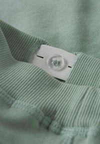 Närbild av ljusgrön stickad tyg med resårband och genomskinlig knapp sydd genom ett knapphål för justerbar storlek.