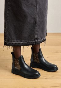 Stivaletti neri in pelle con dettagli in stampa leopardata, punta tonda e suola testurizzata, abbinati a una gonna di jeans nera sfilacciata.