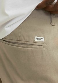 Béžové bavlněné kalhoty s zadní kapsou, na které je bílá etiketa s nápisem "JACK & JONES EST. 1990." Hladká textura a precizní střih.