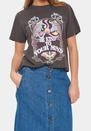 Vrouw draagt een donker T-shirt met kleurrijke vlindervormen en tekst "Be kind to your mind,", gecombineerd met een blauwe spijkerrok met knopen aan de voorkant.