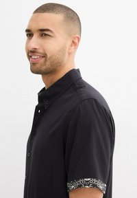 Camicetta nera a maniche corte con rifiniture a strisce bianche e nere sui polsini, apertura con bottoni e tessuto morbido.